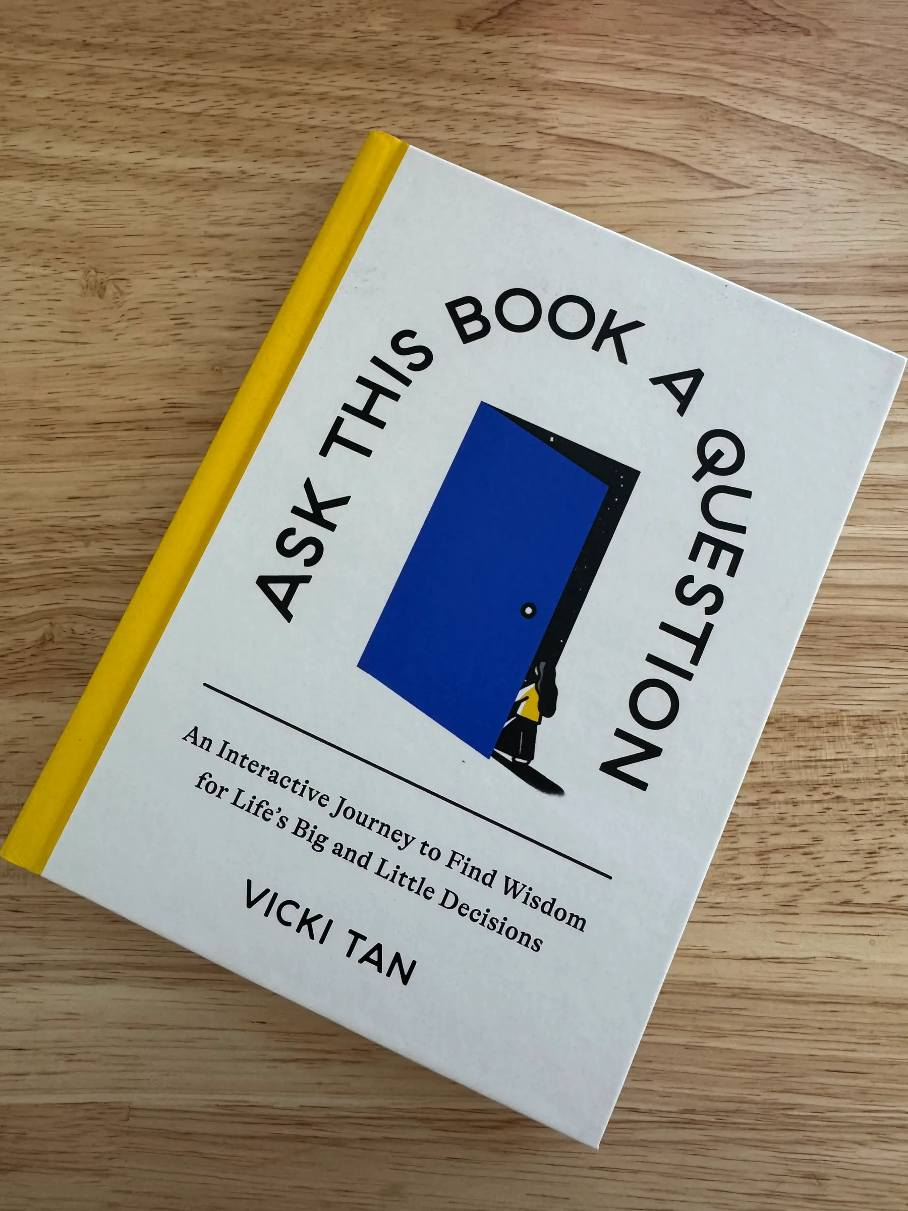 Ask This Book a Question by Vicki Tan lying on a wooden table—a white hardback with a yellow spine, a blue door on the cover