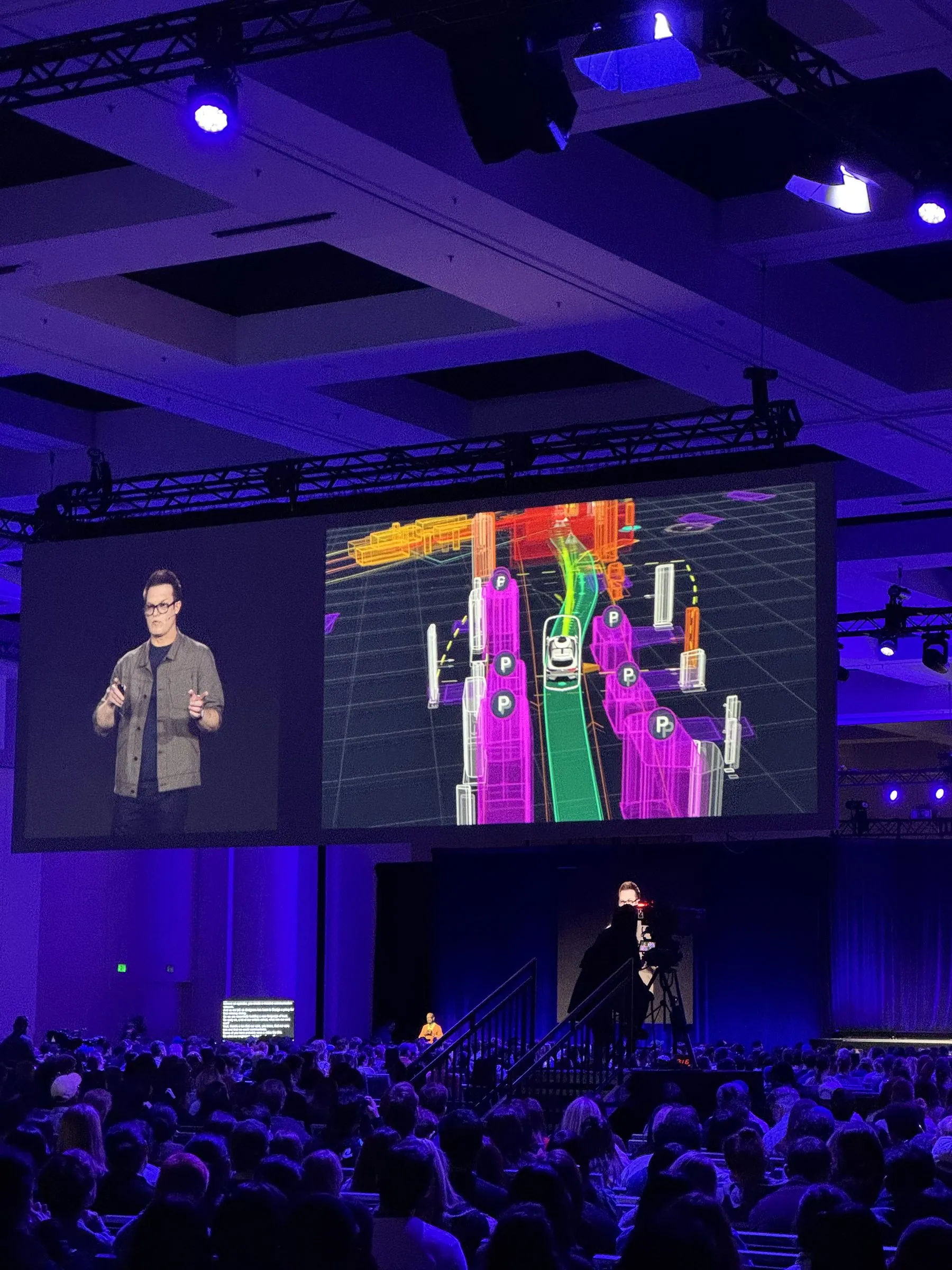 A speaker on the main stage beside a large screen showing a neon wireframe visualization of a self-driving car's view of an intersection
